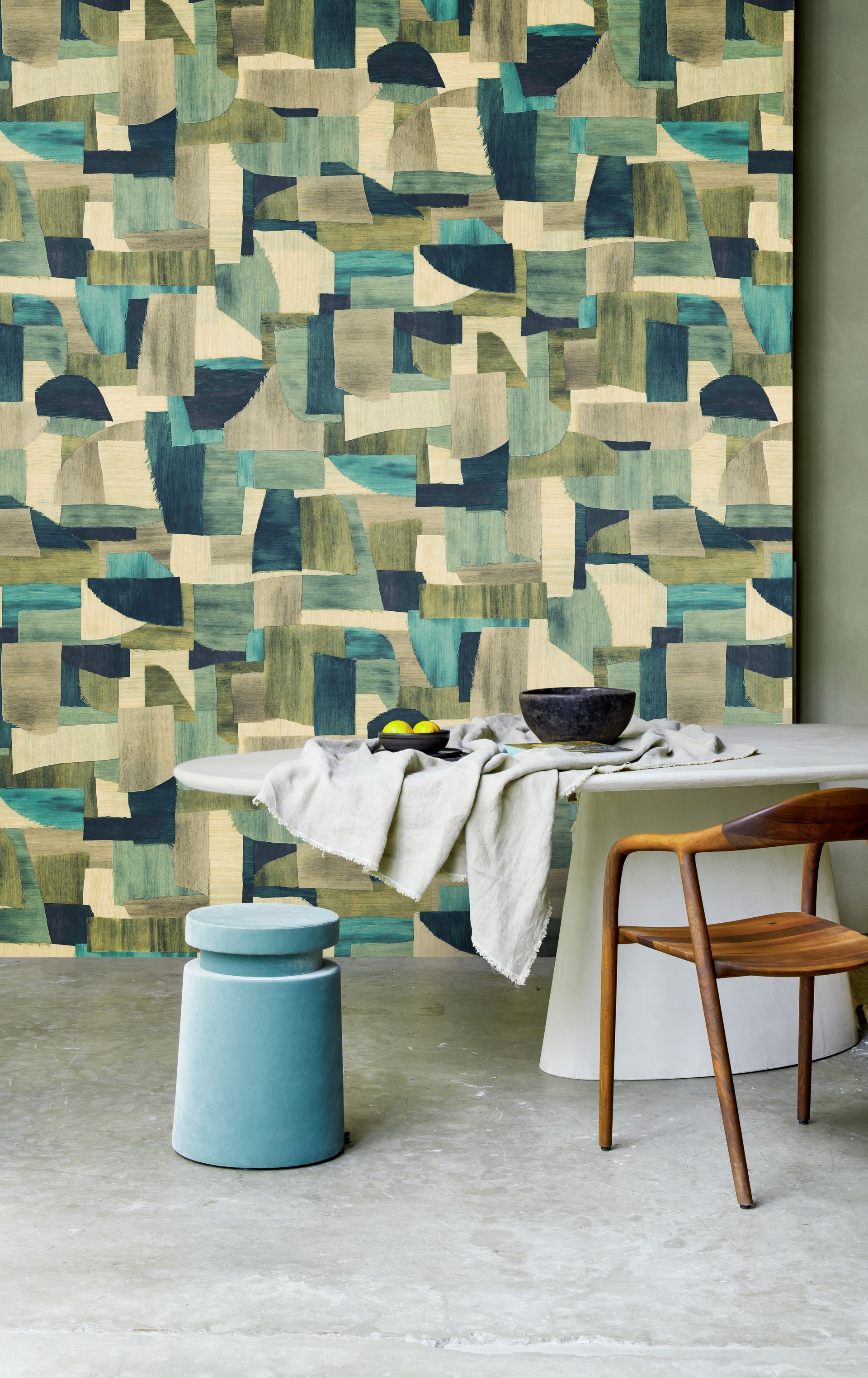 Parete con carta da parati astratta nei toni del blu, verde e beige, dietro un tavolo bianco apparecchiato per la cena con sgabello azzurro e sedia in legno.