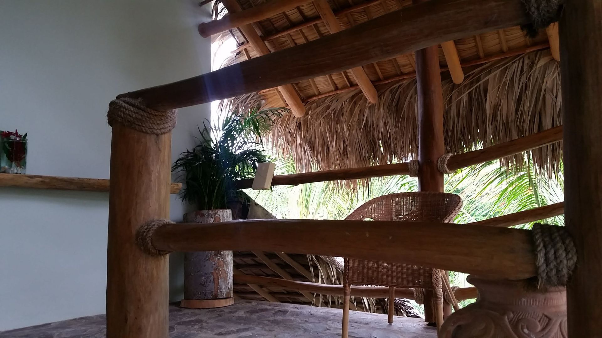 Wooden railing with rope accents, view of a thatched roof and a chair in an outdoor setting.