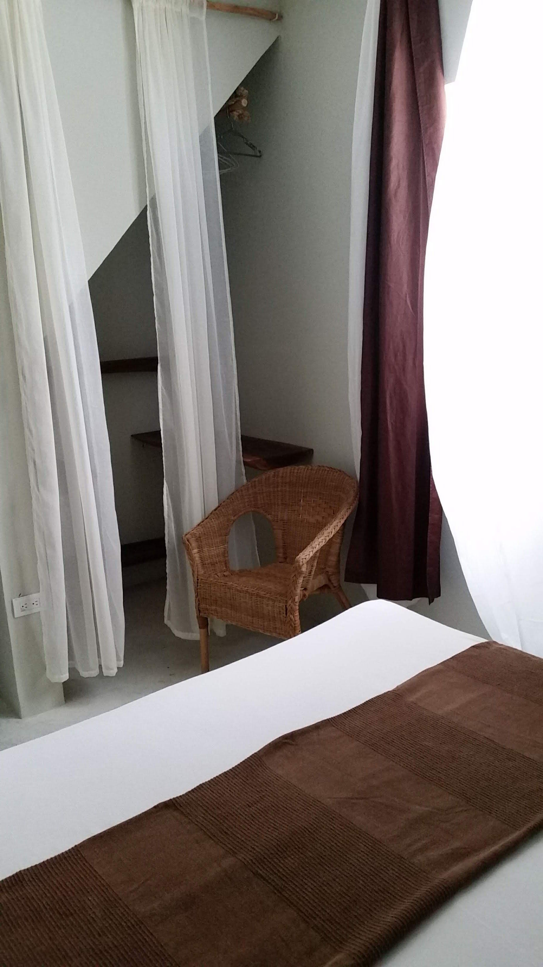 Bedroom with bed, wicker chair, white curtains, and brown drapes. Closet with shelves visible.