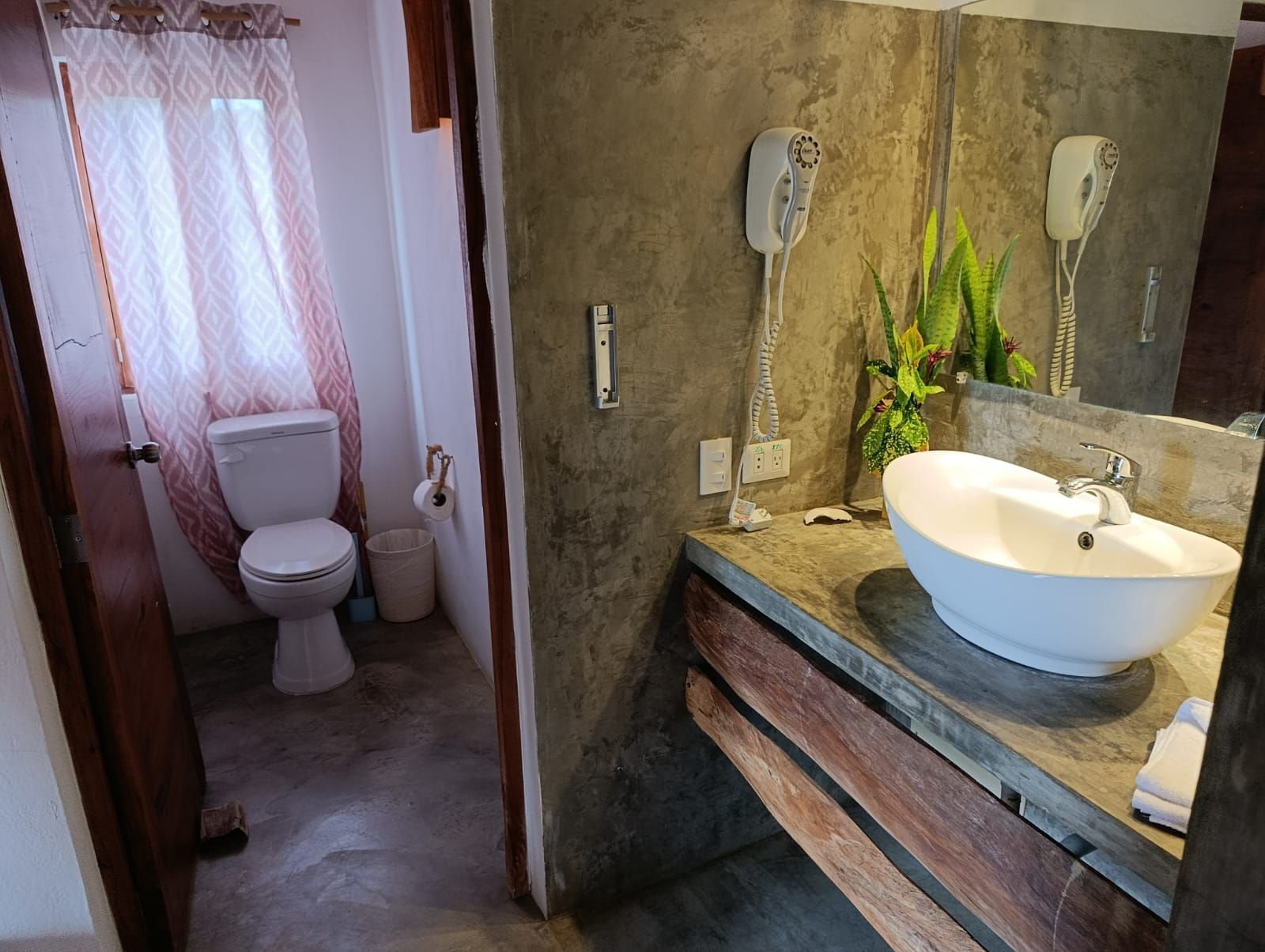 Bathroom with a toilet, sink, and mirror. Concrete walls and wooden accents create a rustic look.