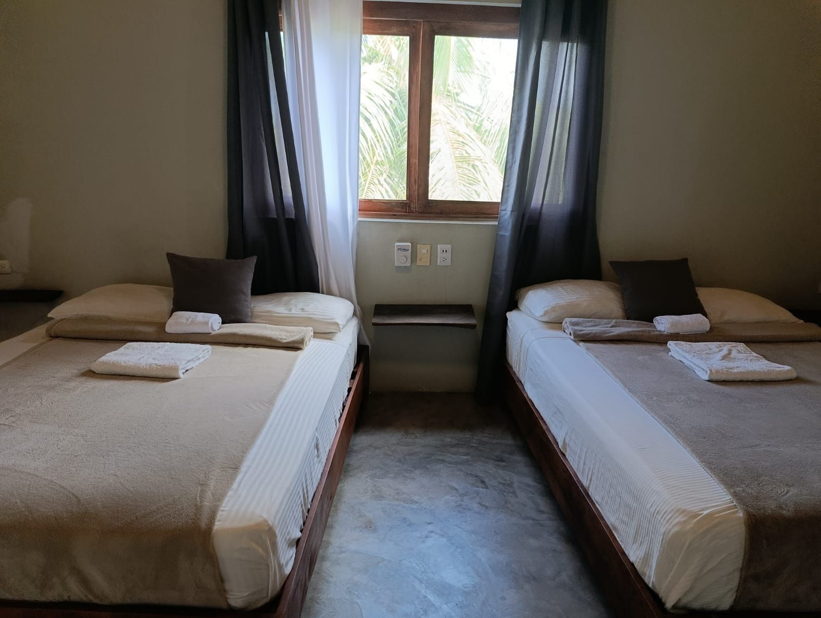 Two beds in a room, with a window between them. Beige bedding, dark brown pillows.