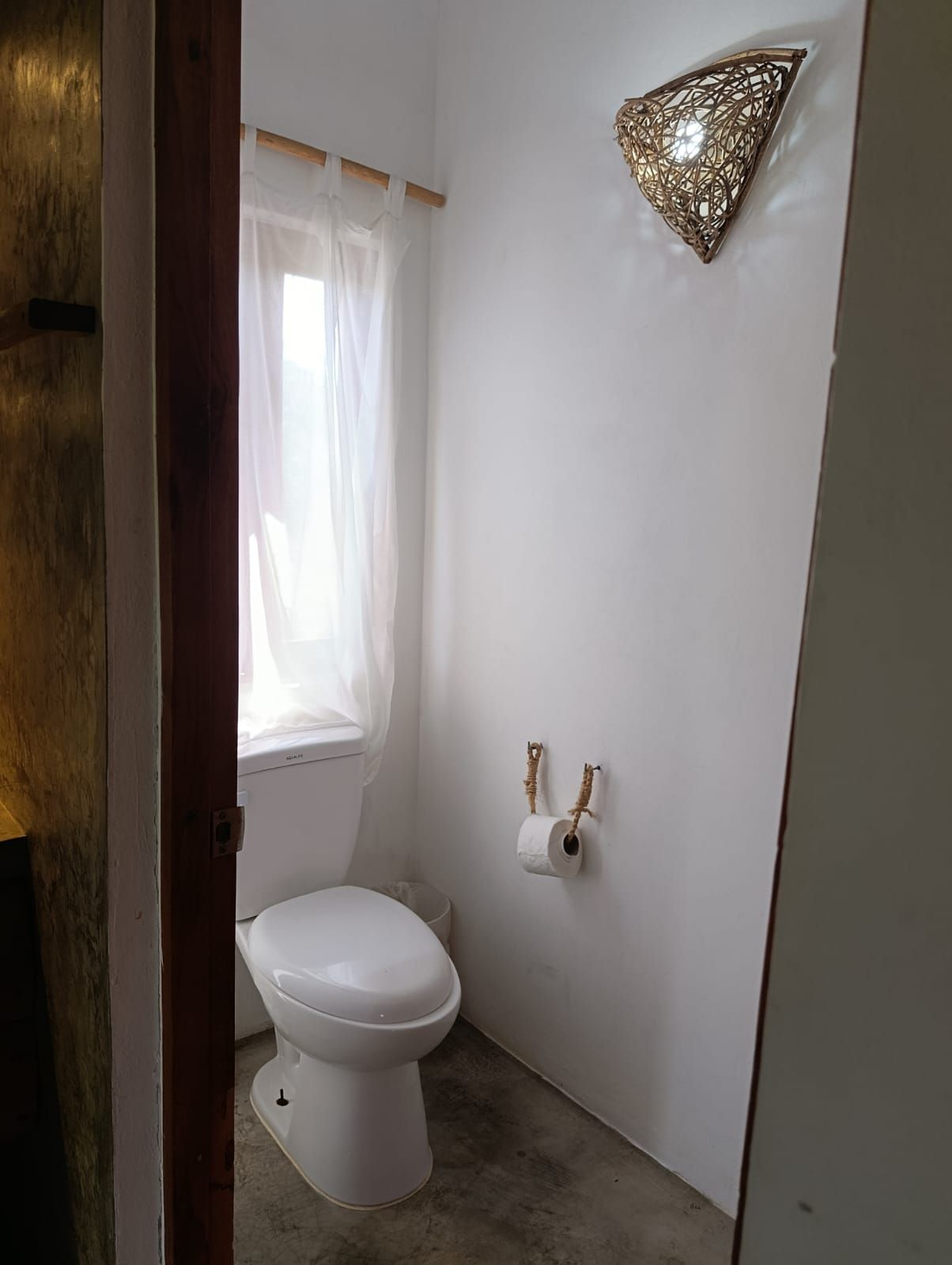 Small, white bathroom with a toilet, window with sheer curtain, and decorative wall sconce.