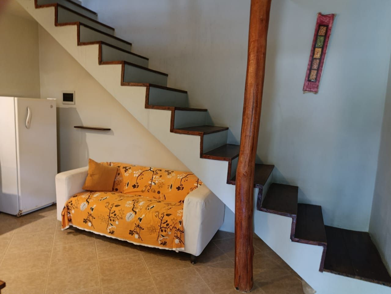 Small living area with staircase, white sofa with yellow cover, refrigerator, and decorative wall art.