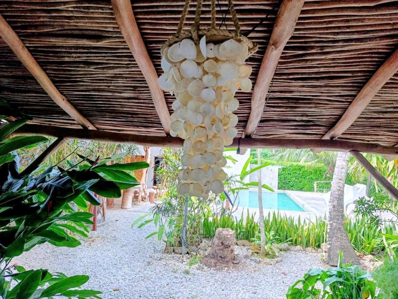 Shell chandelier hangs under a wooden-beamed roof, overlooking a garden and pool.