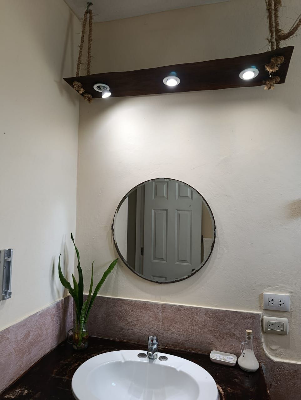 Bathroom with round mirror, hanging light fixture, and sink. Dark countertop, off-white walls.
