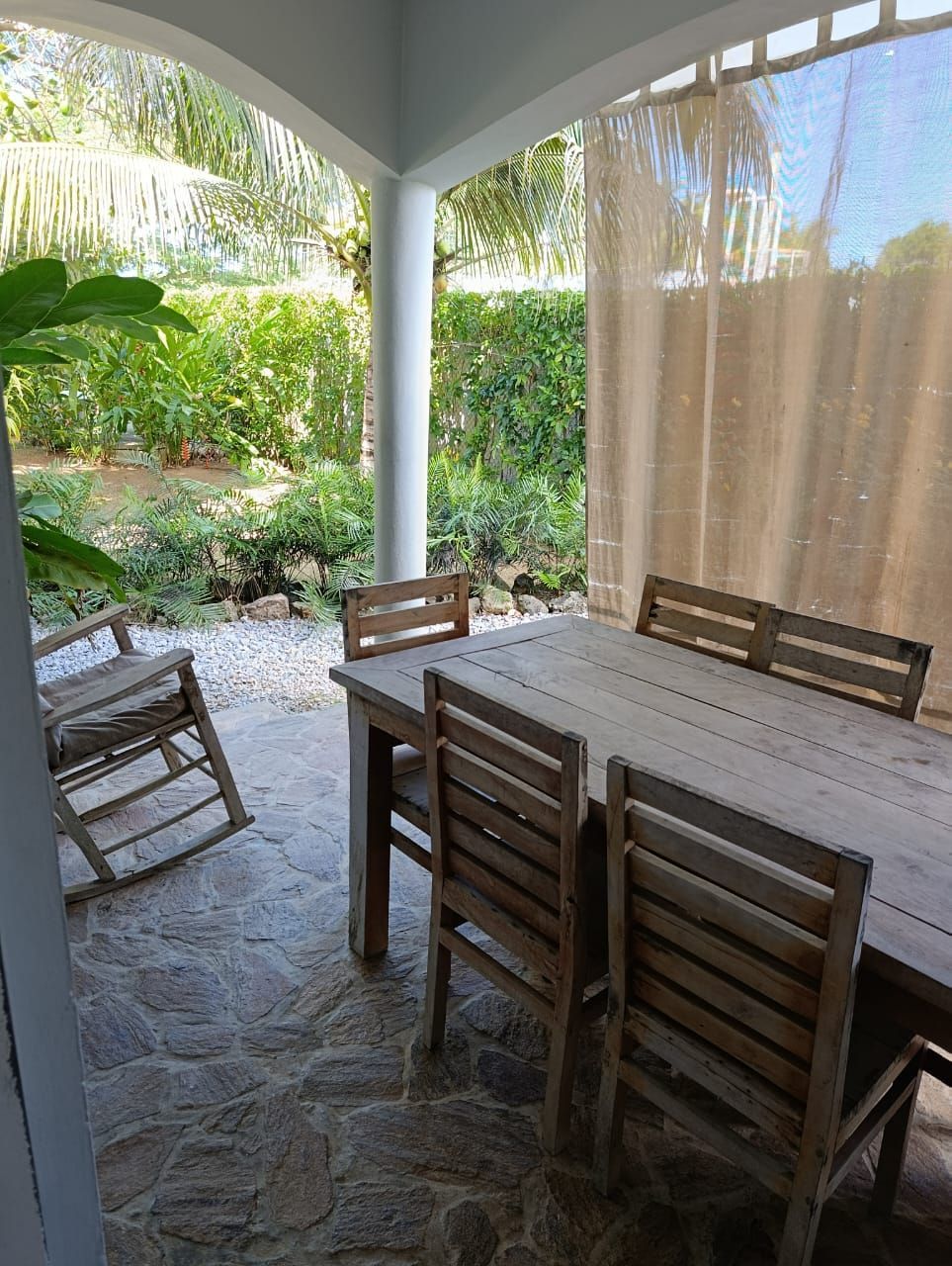 Covered outdoor dining area with wooden table, chairs, and rocking chair, overlooking a garden.