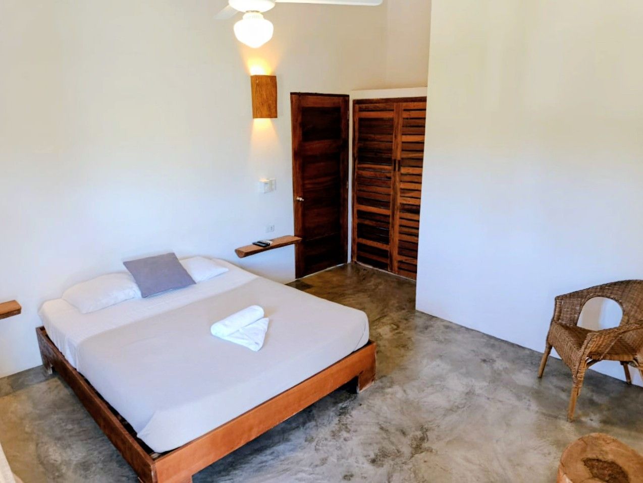 Bedroom with a bed, wooden furniture, and a woven chair. The walls are white and the floor is concrete.
