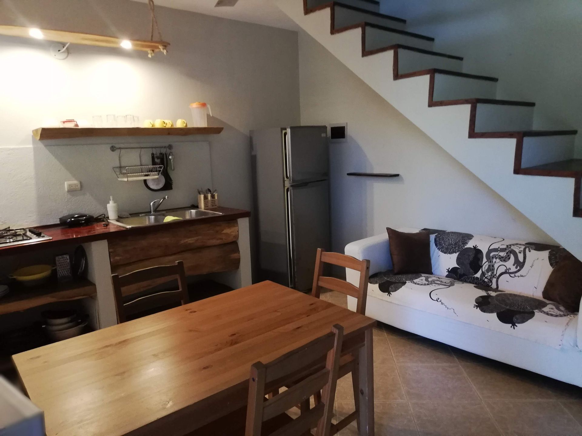 Kitchen and living area with wooden table and chairs, sofa, stairs, and refrigerator.