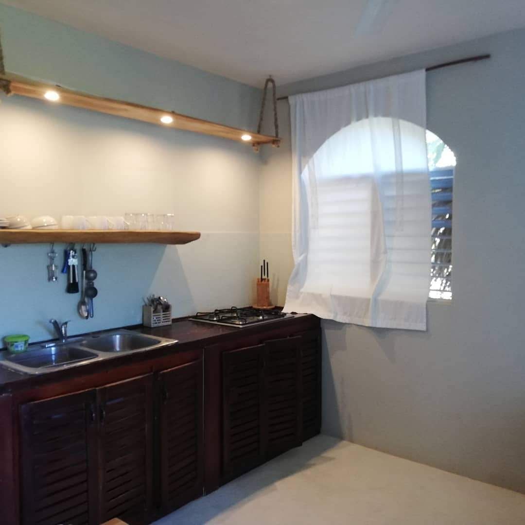Small kitchen with dark wooden cabinets, double sink, stove, and a window with white curtains.