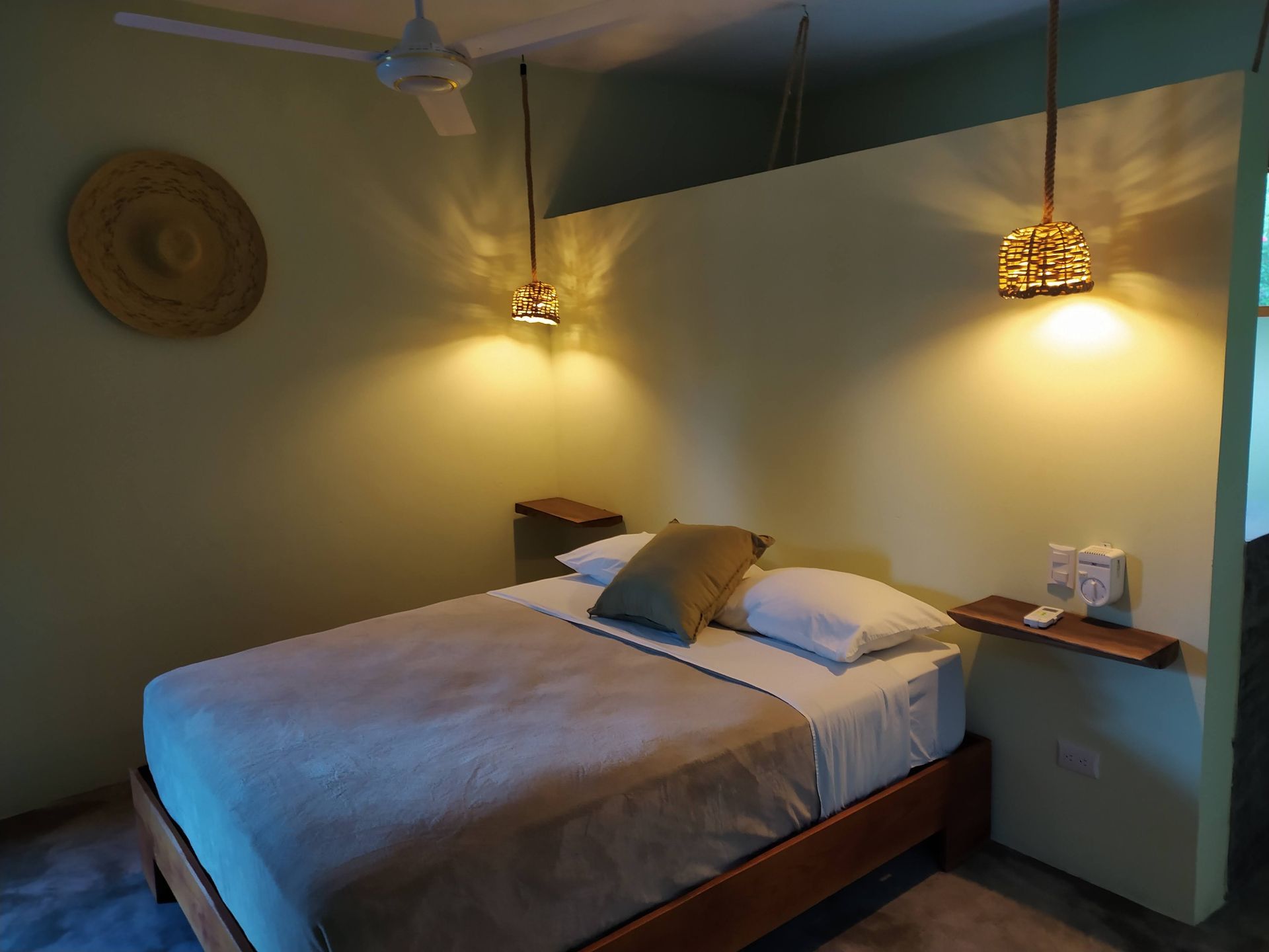 Bedroom with a bed, two lamps, and a woven wall decoration. Pale green walls and wooden accents.
