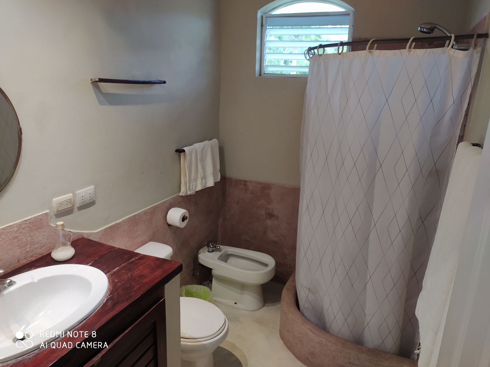 Bathroom with sink, toilet, bidet, and shower. Tan and white tones, small window.