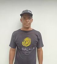 Man wearing a cap and black t-shirt with a yellow design, standing against a white wall.