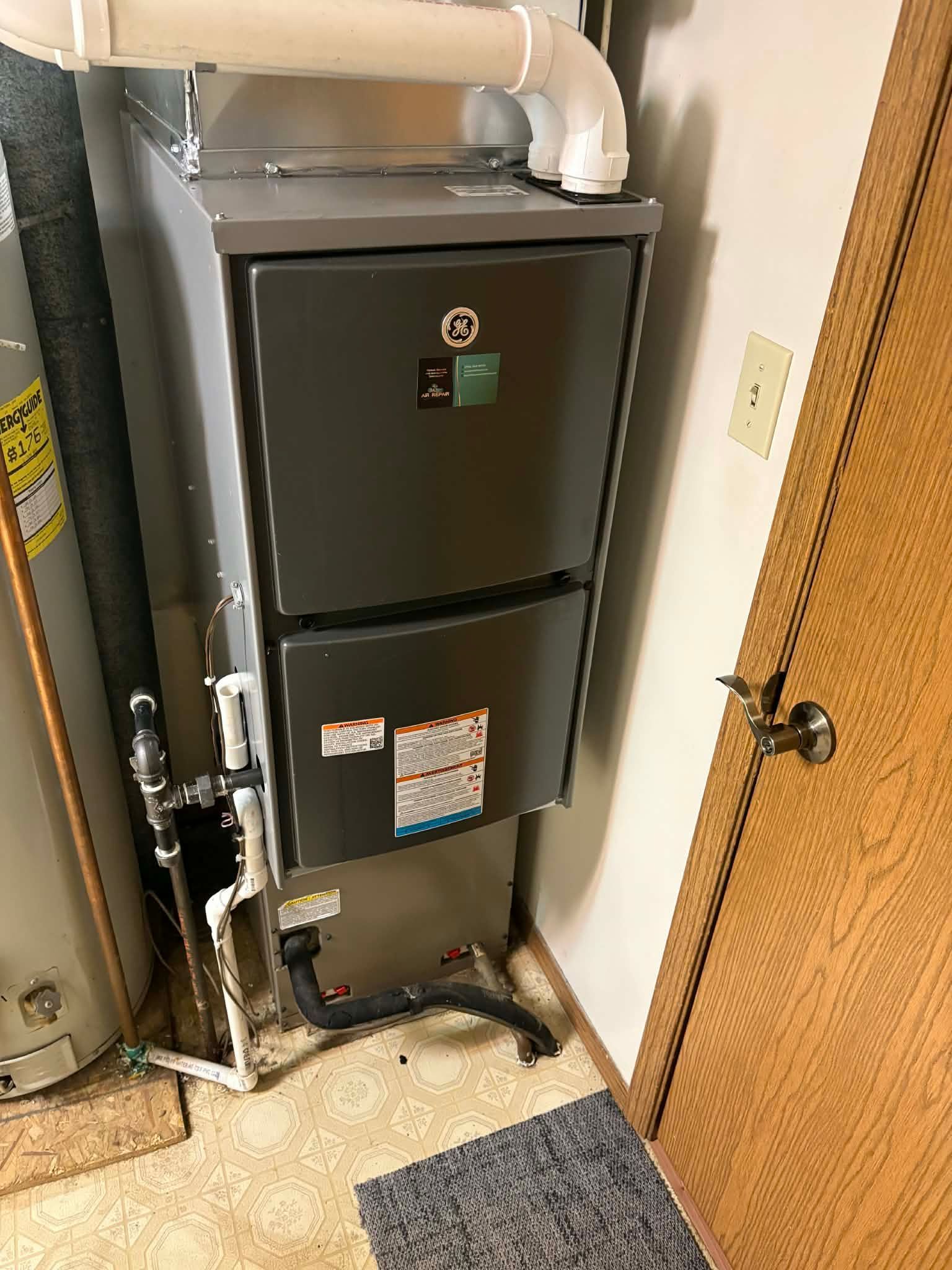 A grey gas furnace with white PVC venting pipes, standing on a carpeted floor next to a water heater and wooden door.