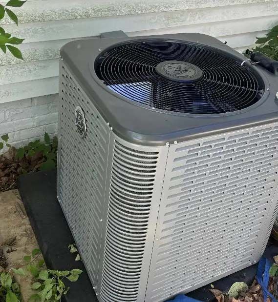 Outdoor air conditioner unit beside a house wall, with a black fan on top and silver grille sides