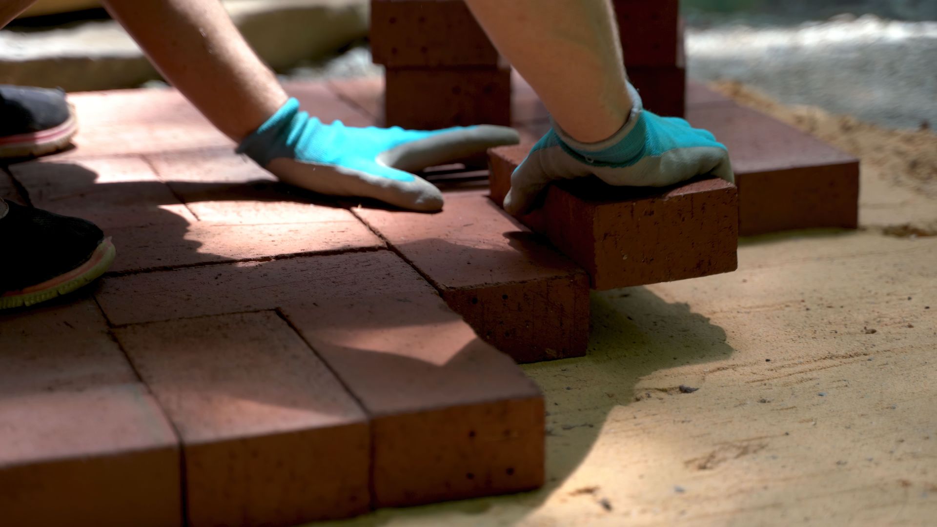 Hands in blue gloves laying red bricks on sandy ground