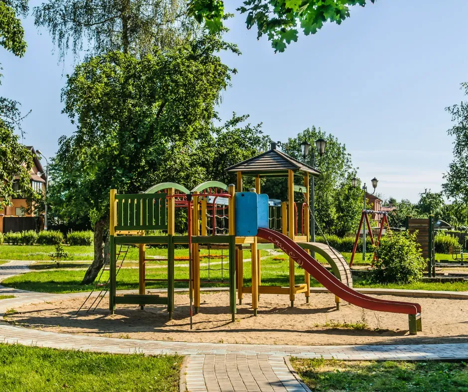 Playground with a slide, climbing structure, and swing set, surrounded by grass and trees.