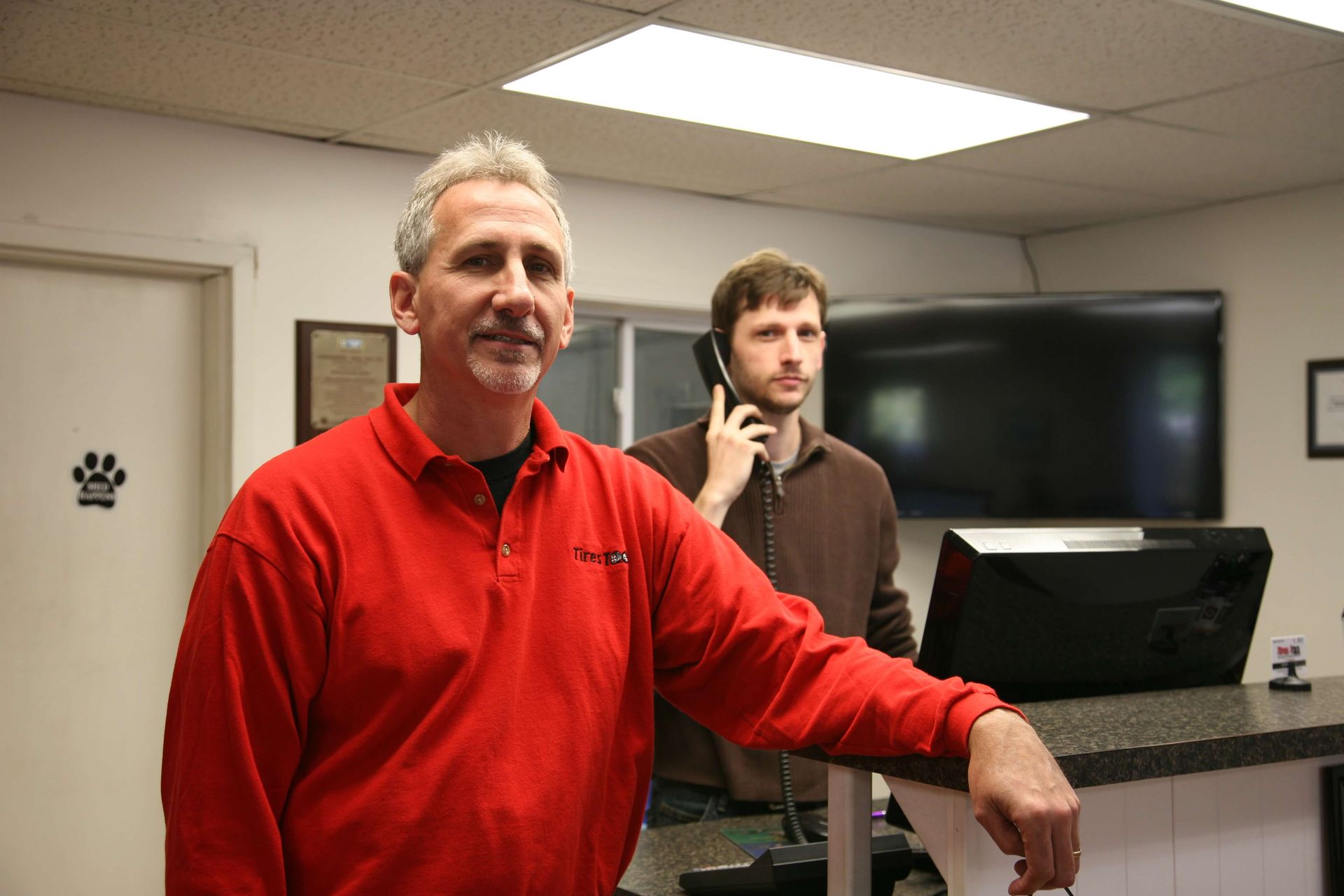 Two people at a reception desk in an office, one in a red shirt leaning on the counter.
