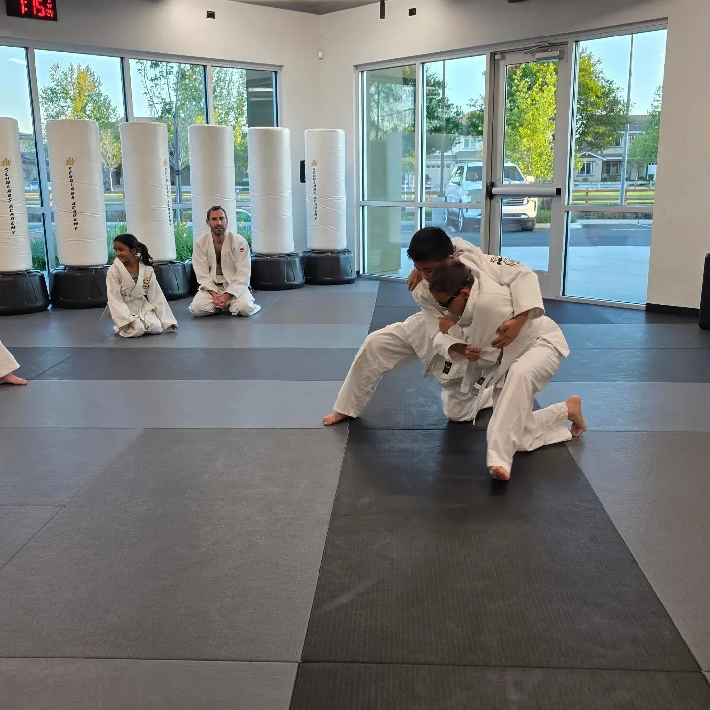 Children practicing a supervised judo grappling technique on padded mats at Scholars Academy in Brentwood, CA