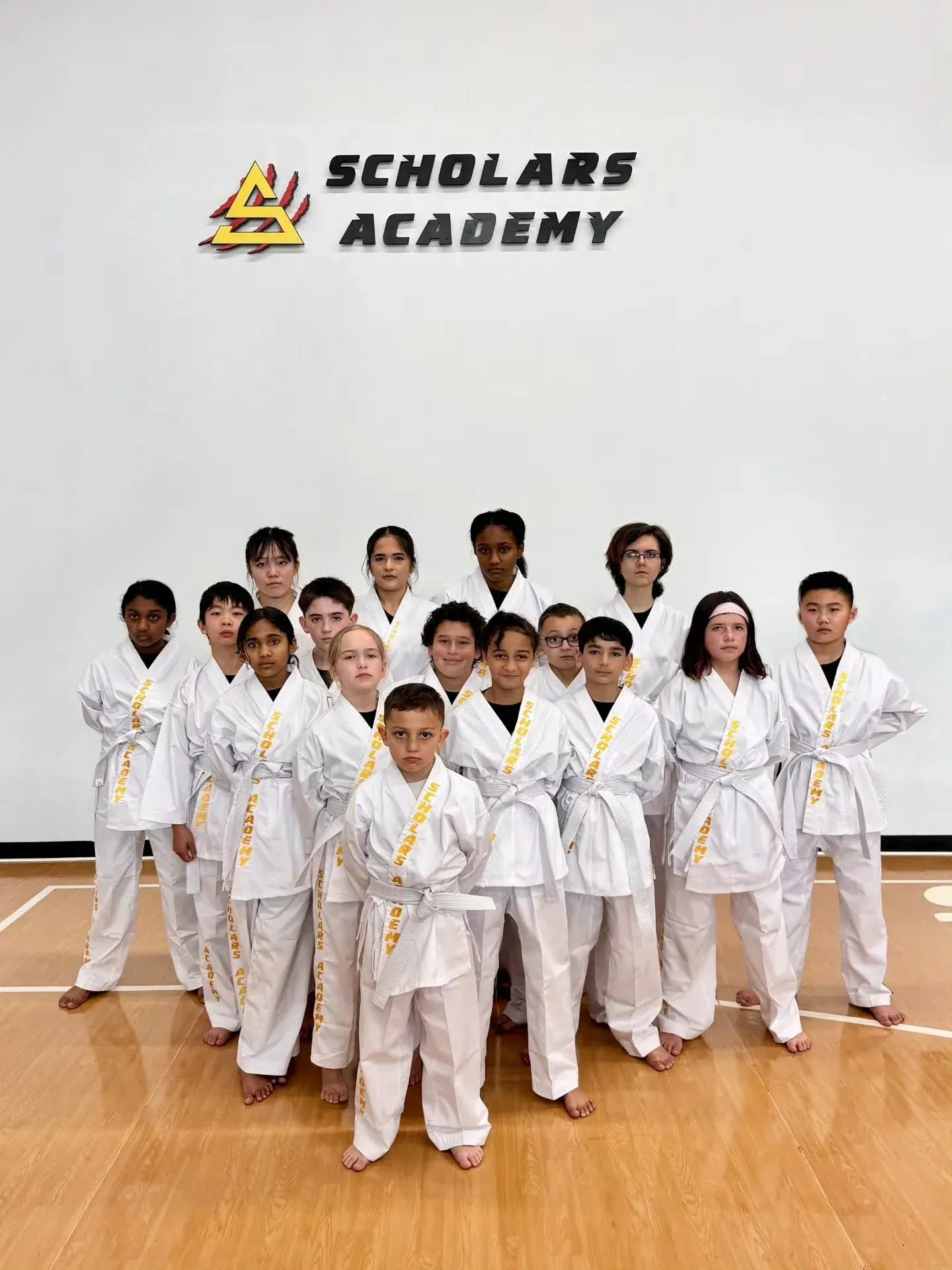 Karate students in white uniforms posing in a dojo with a Scholars Academy logo