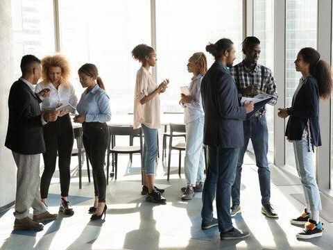 Diverse group of professionals standing and talking in a sunlit office space with large windows.