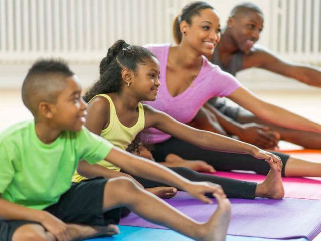 Four people sitting on colorful yoga mats on the floor, stretching their arms forward toward their feet in a fitness studio.