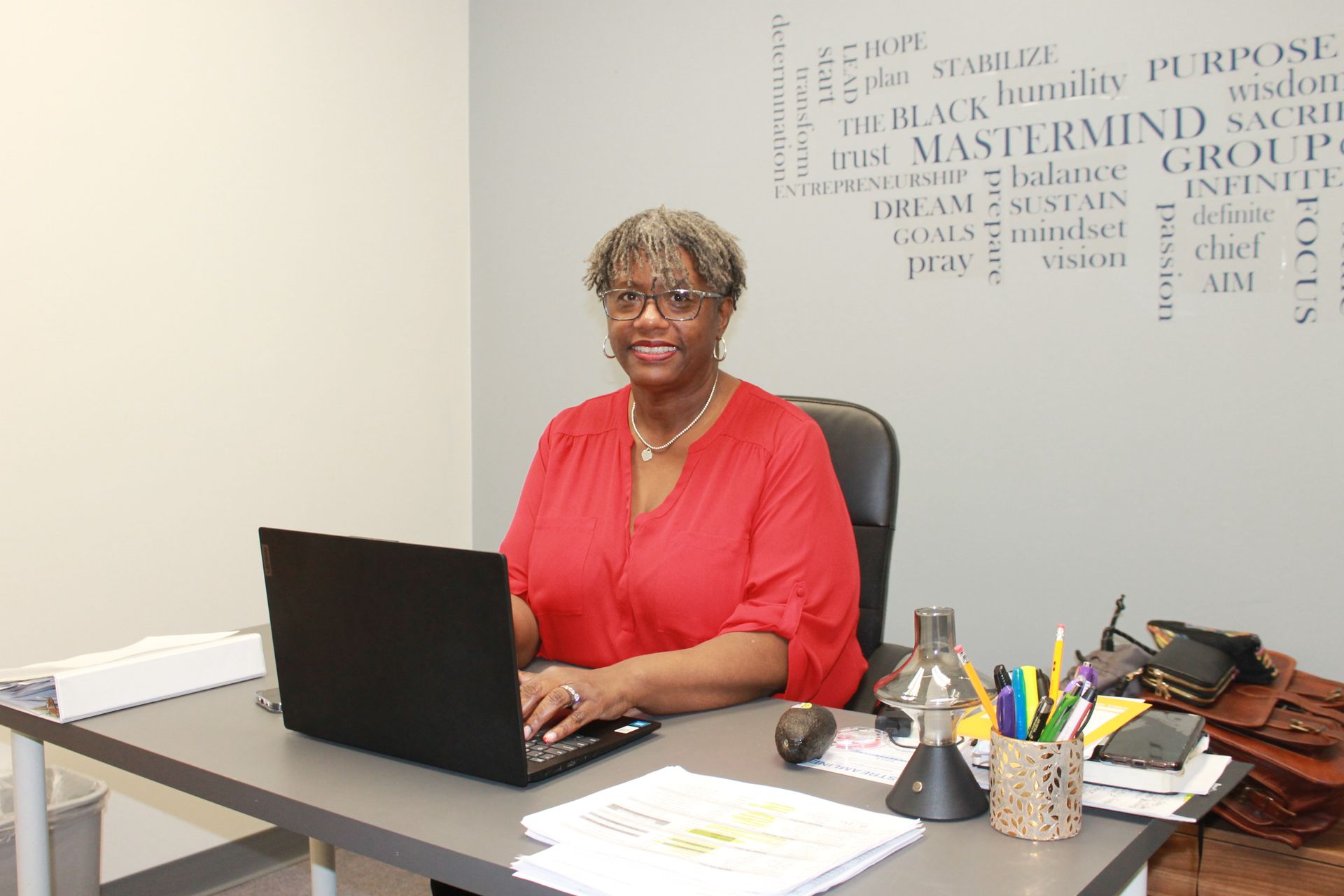 A person wearing a red shirt sits at a desk typing on a laptop in an office with a word-collage wall decal.