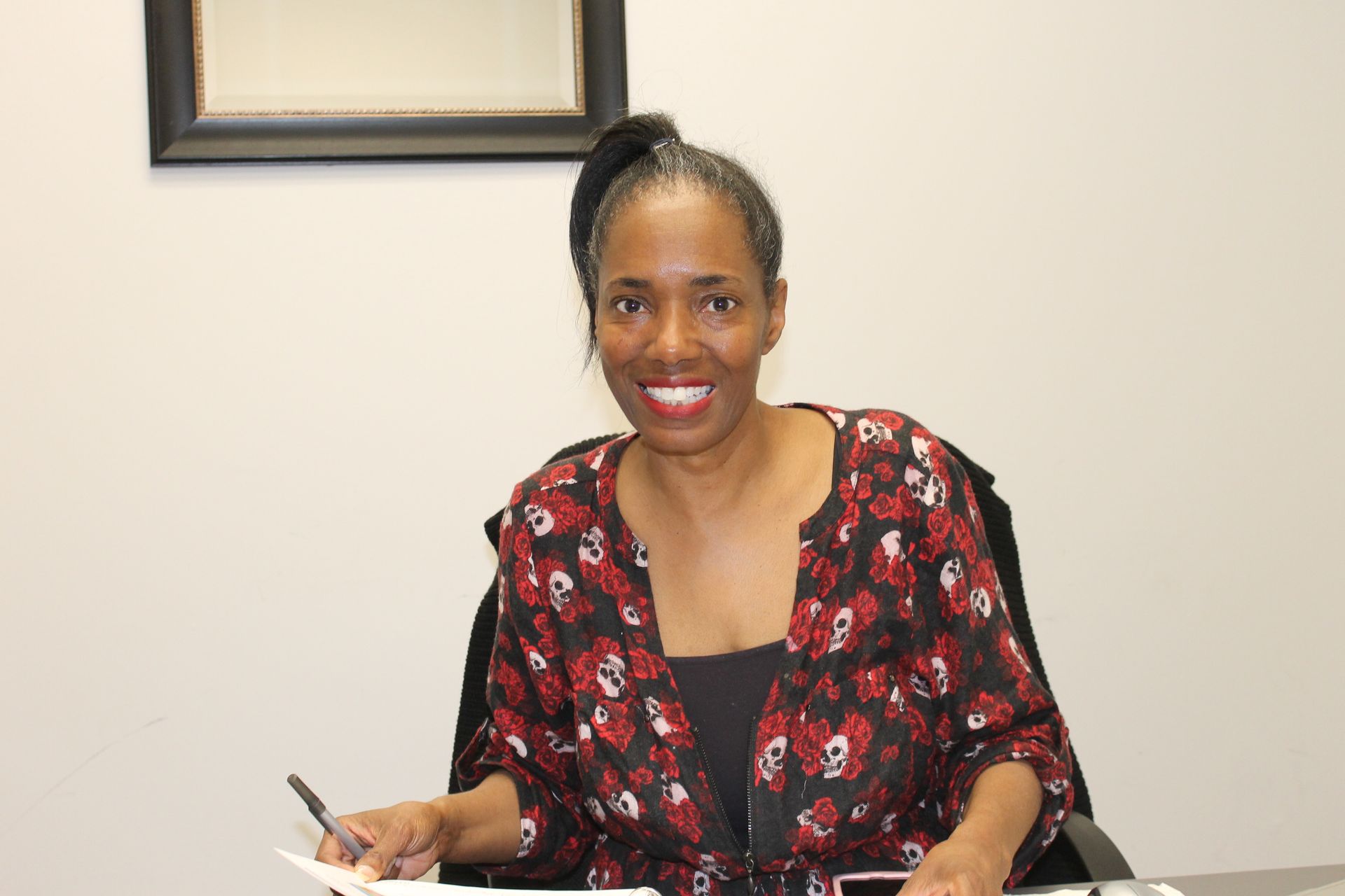 A person with their hair in a high bun smiles while holding a pen at a desk in front of a white wall.