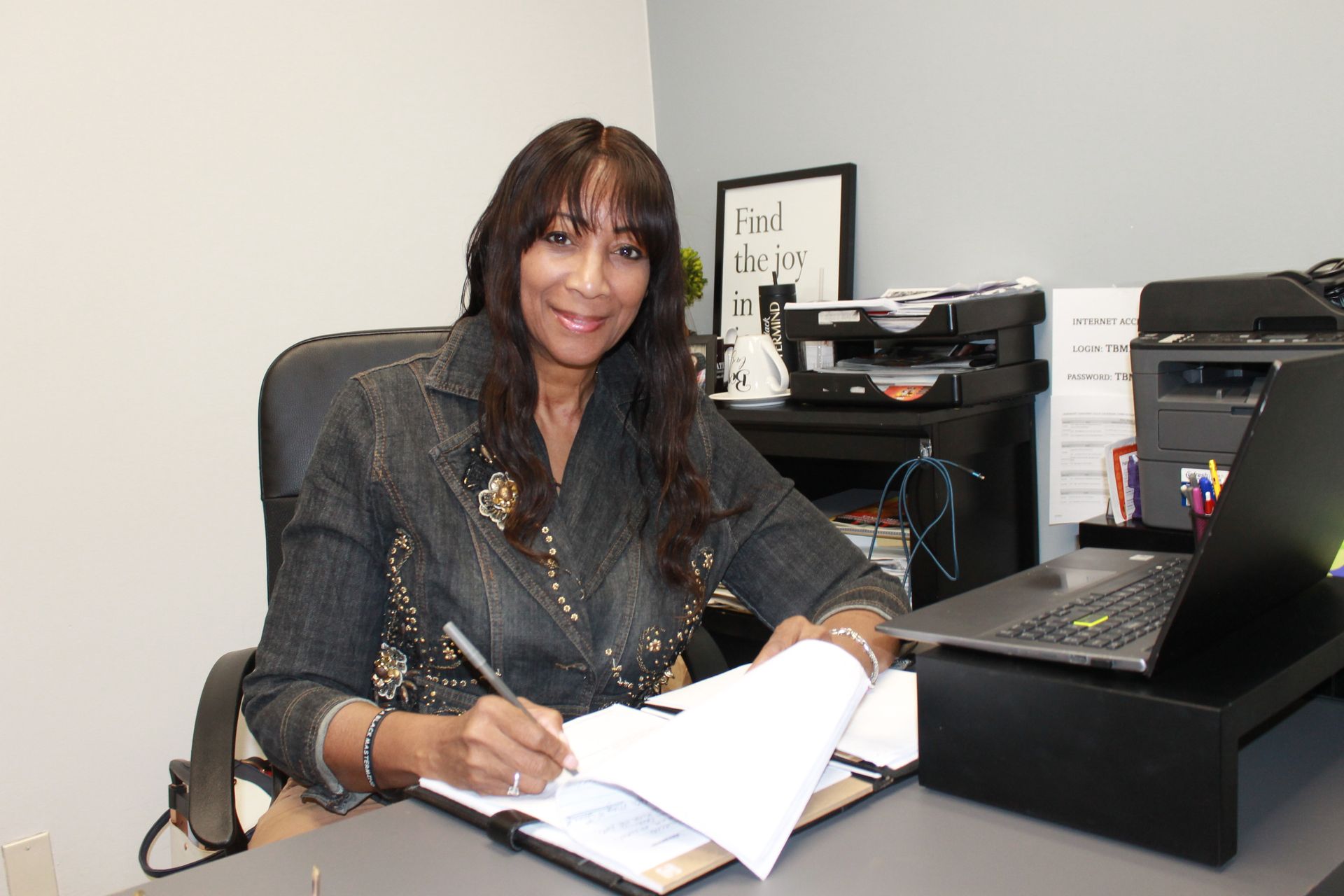 A professional seated at a desk writing on a document with a laptop and printer in a neutral office setting.