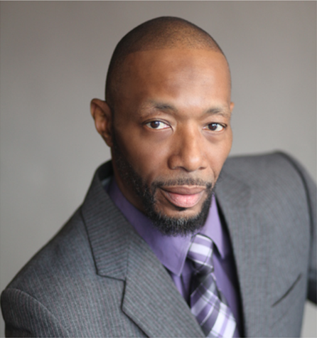 Man in gray suit and purple shirt, looking directly at the viewer with a neutral expression.