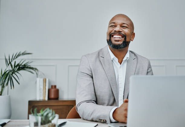 Smiling man in a light gray suit sits at a desk with a laptop, looking up and to the side.