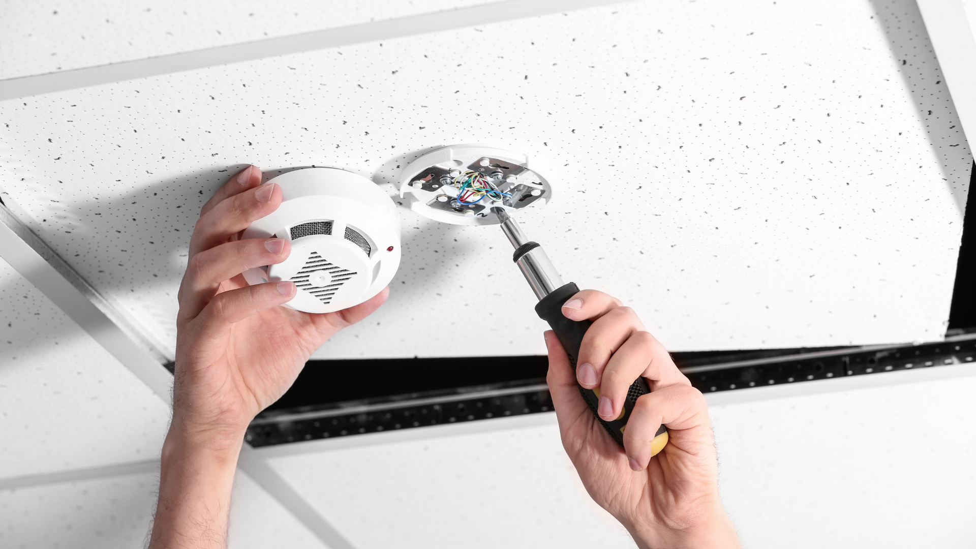 Hands installing a smoke detector on a ceiling tile with a screwdriver