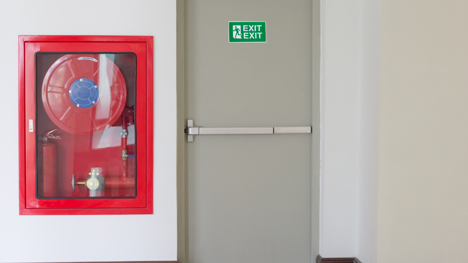 Red fire hose cabinet beside a gray emergency exit door with green exit sign above it