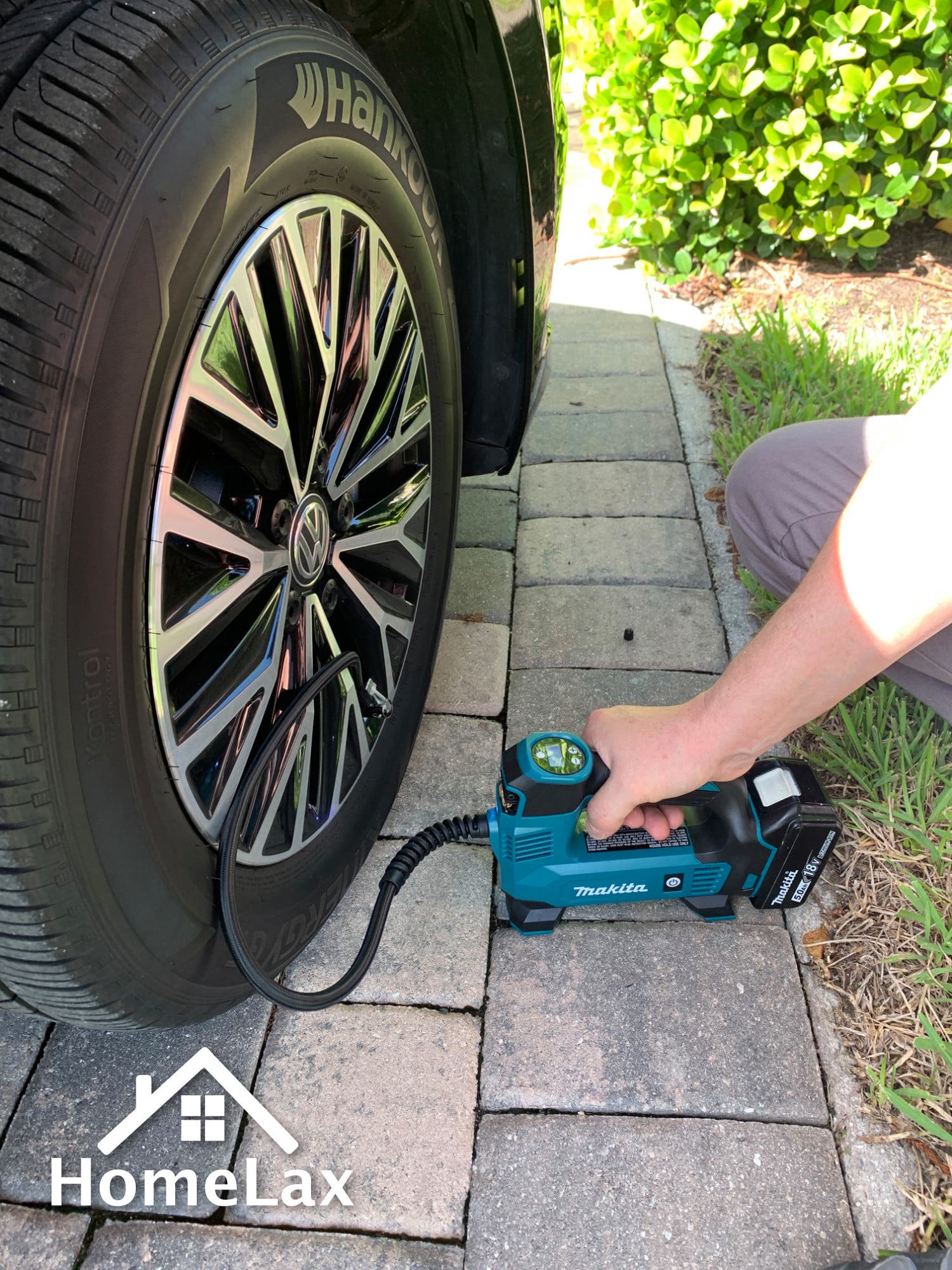 A person using a cordless teal-colored air compressor to inflate a car tire on a paved driveway next to a lawn.