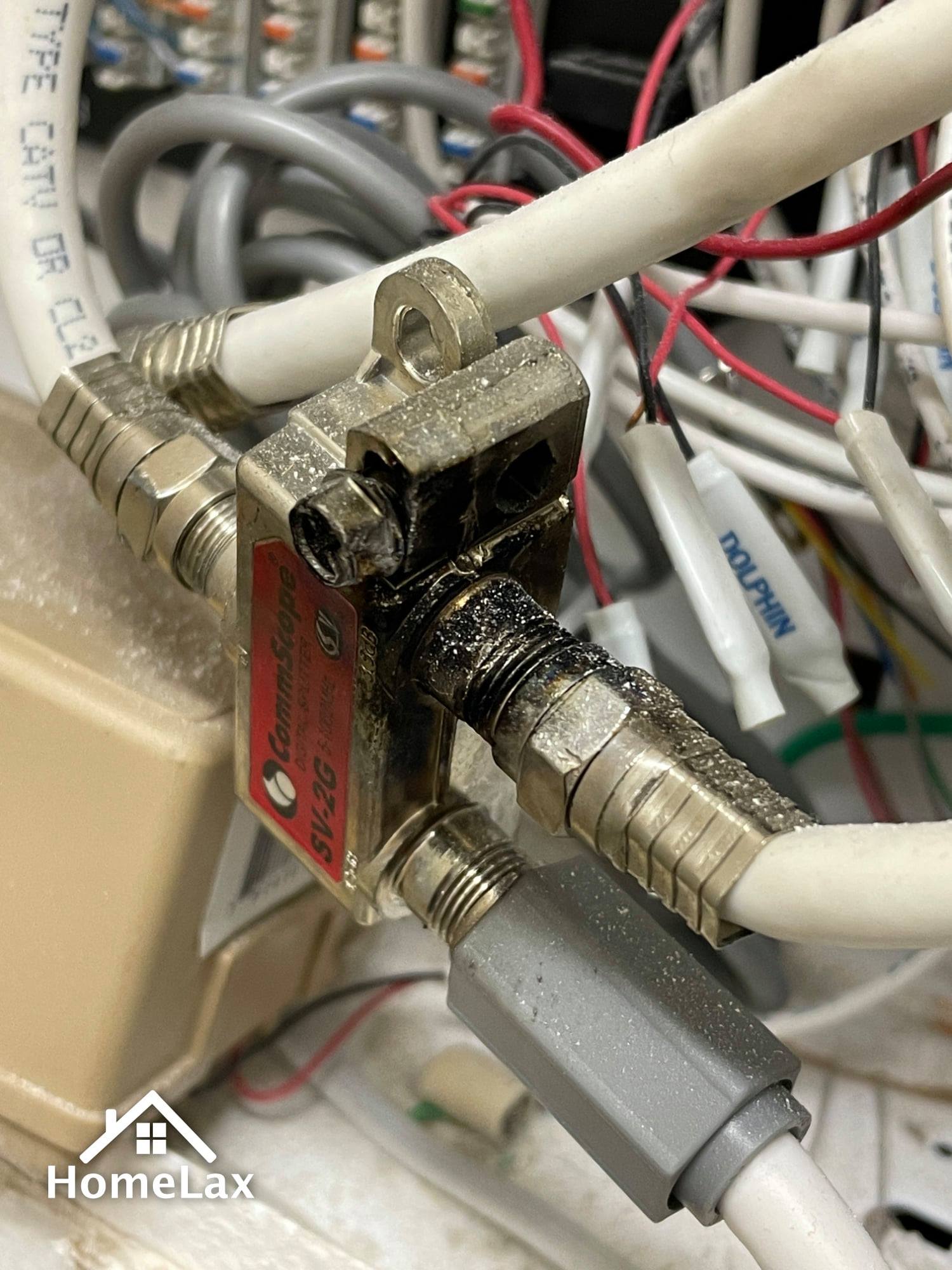 A metallic coaxial cable splitter mounted on a wall with several white cables connected to it in a utility space.