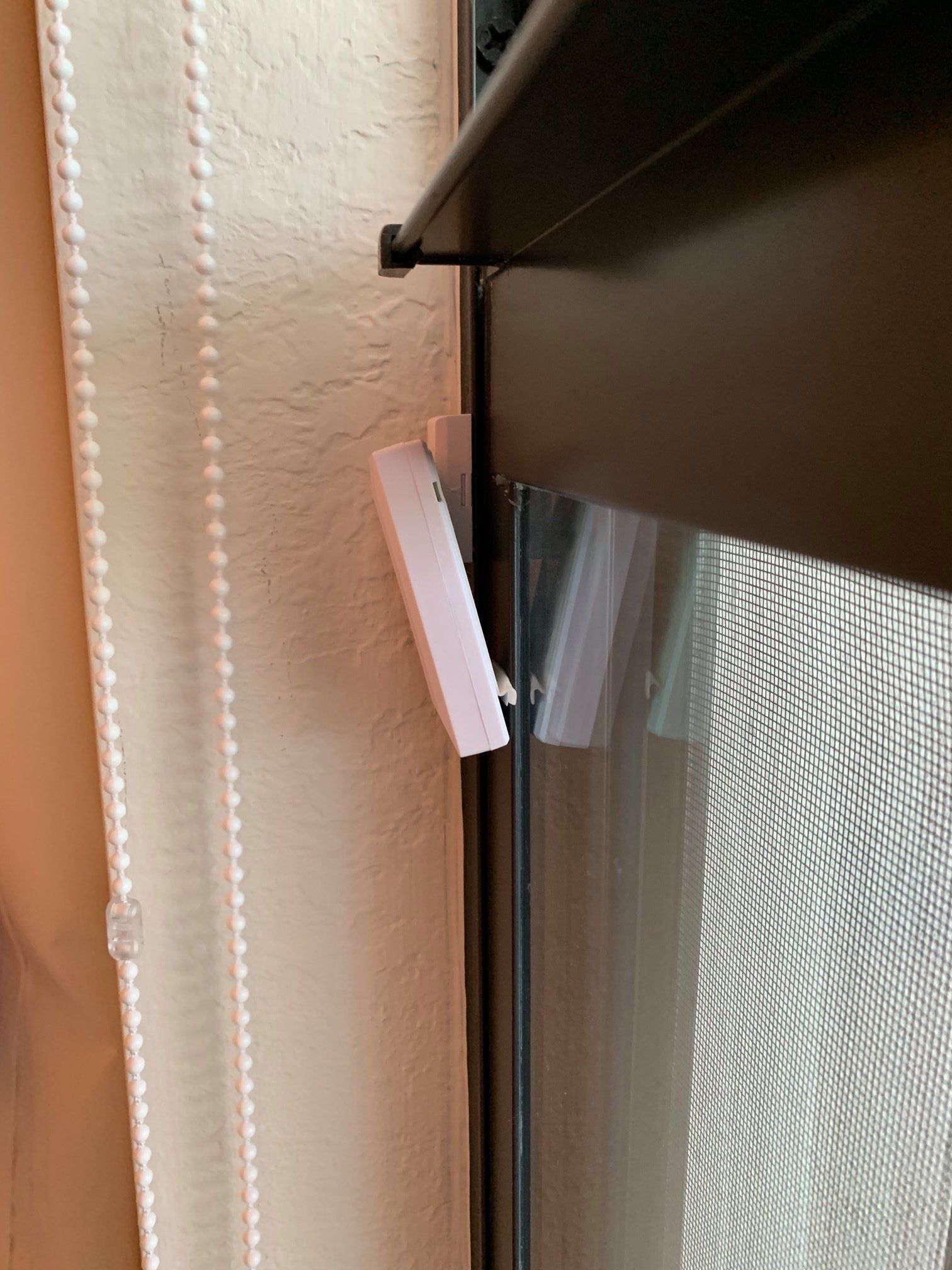 A white rectangular security sensor attached to a wall next to a dark window frame with a beaded curtain cord nearby.