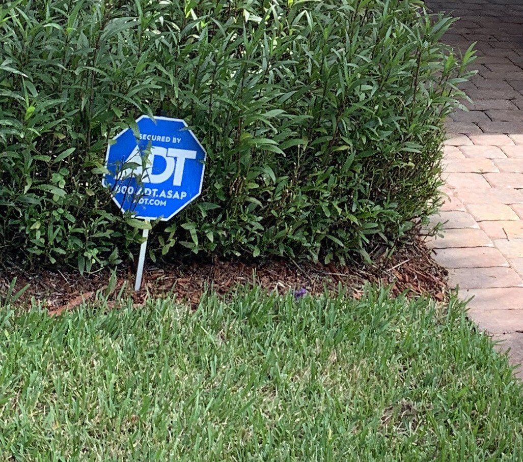 A blue octagonal ADT security sign stands in a green hedge next to a brick walkway.