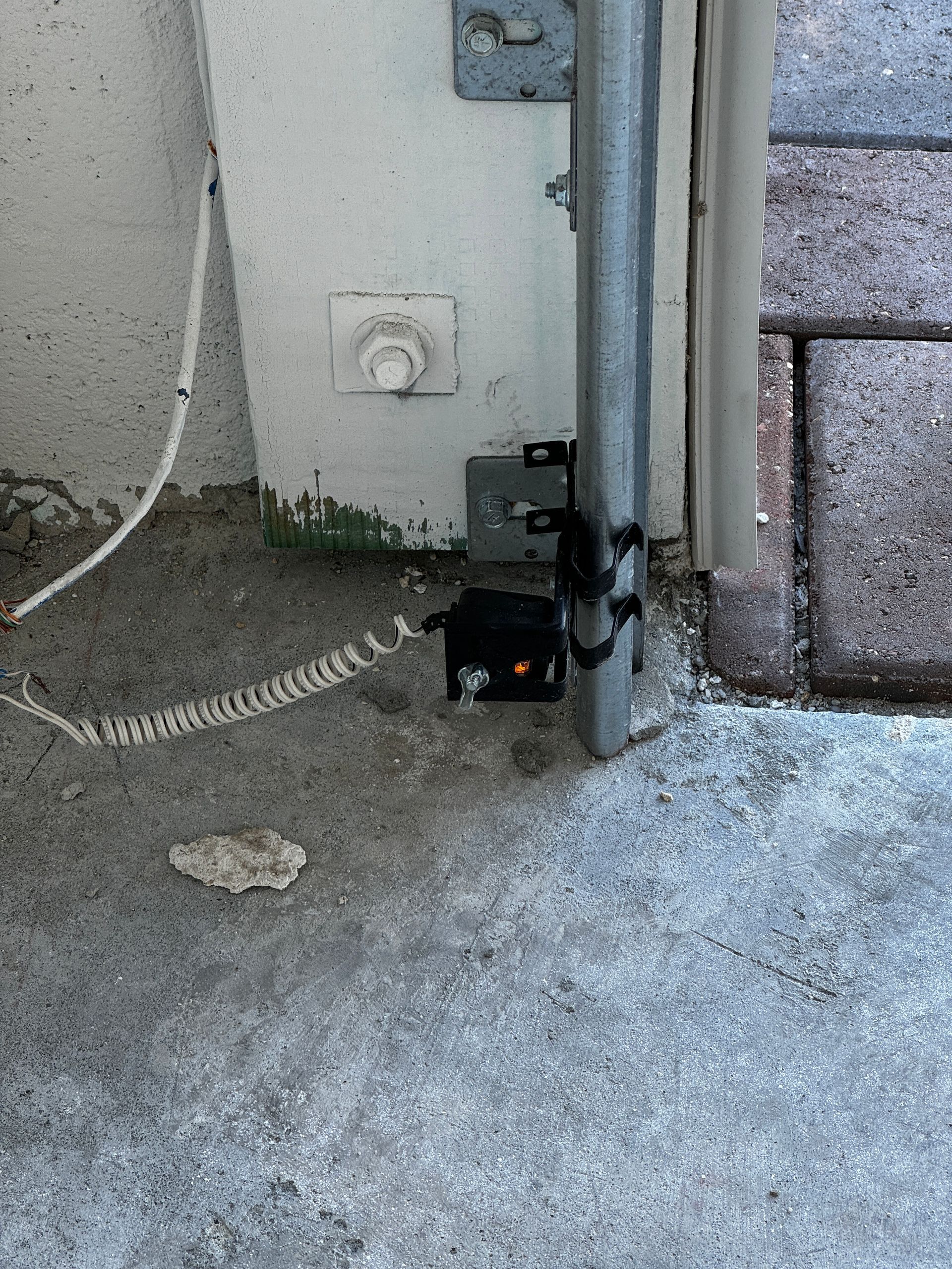 A safety sensor for a garage door opener attached to a metal vertical track against a white wall.