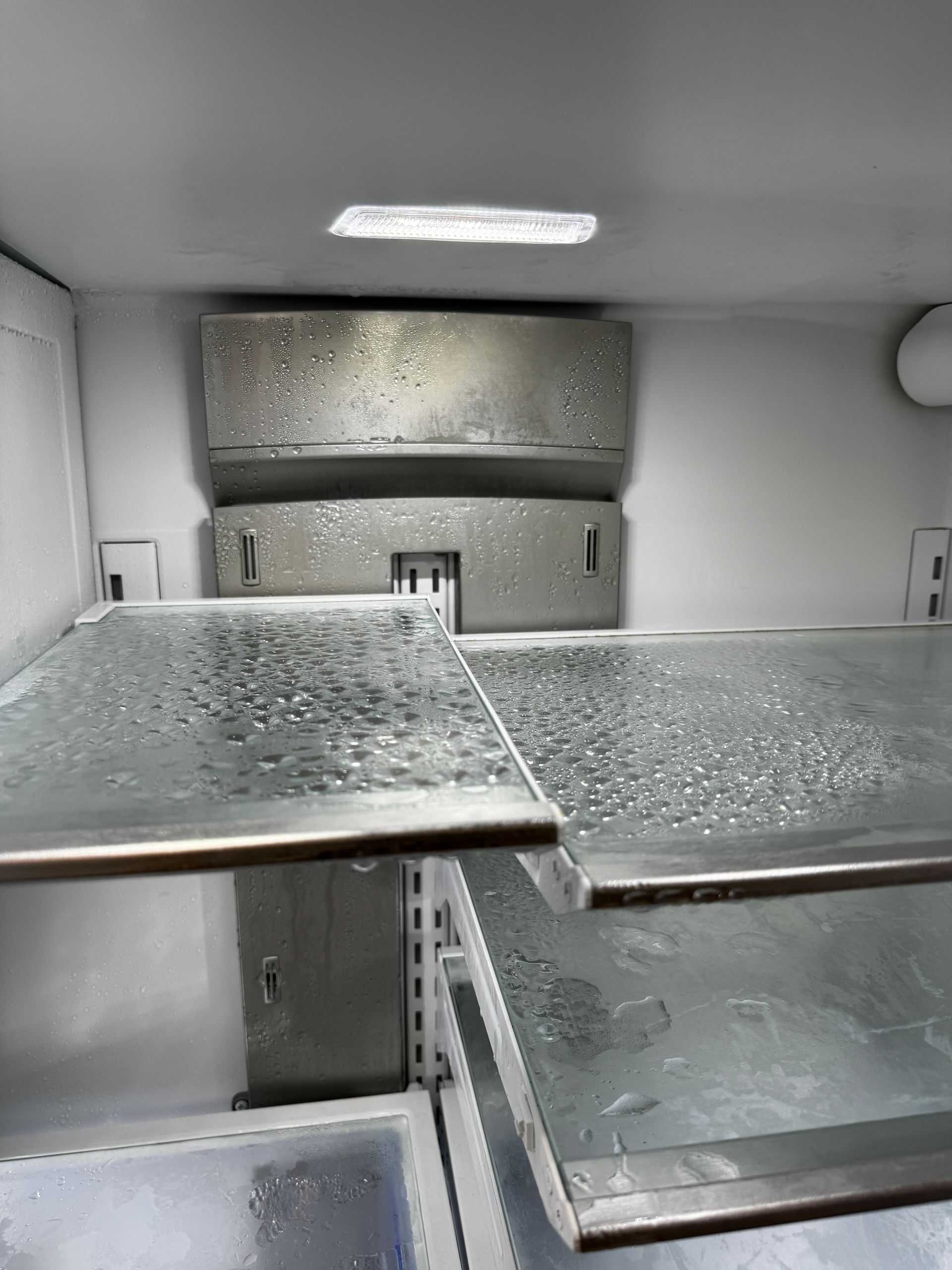 The interior of an empty refrigerator with glass shelves covered in water droplets and condensation.