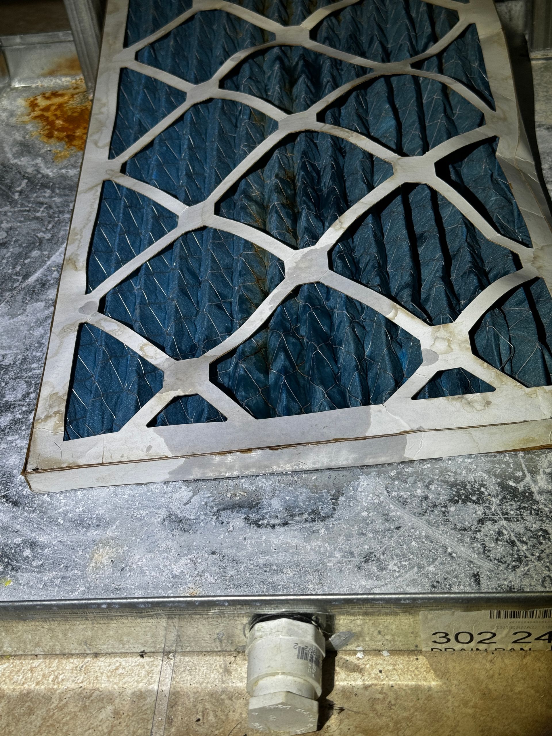 A dirty blue HVAC air filter resting on a metallic unit next to a white PVC drain pipe.