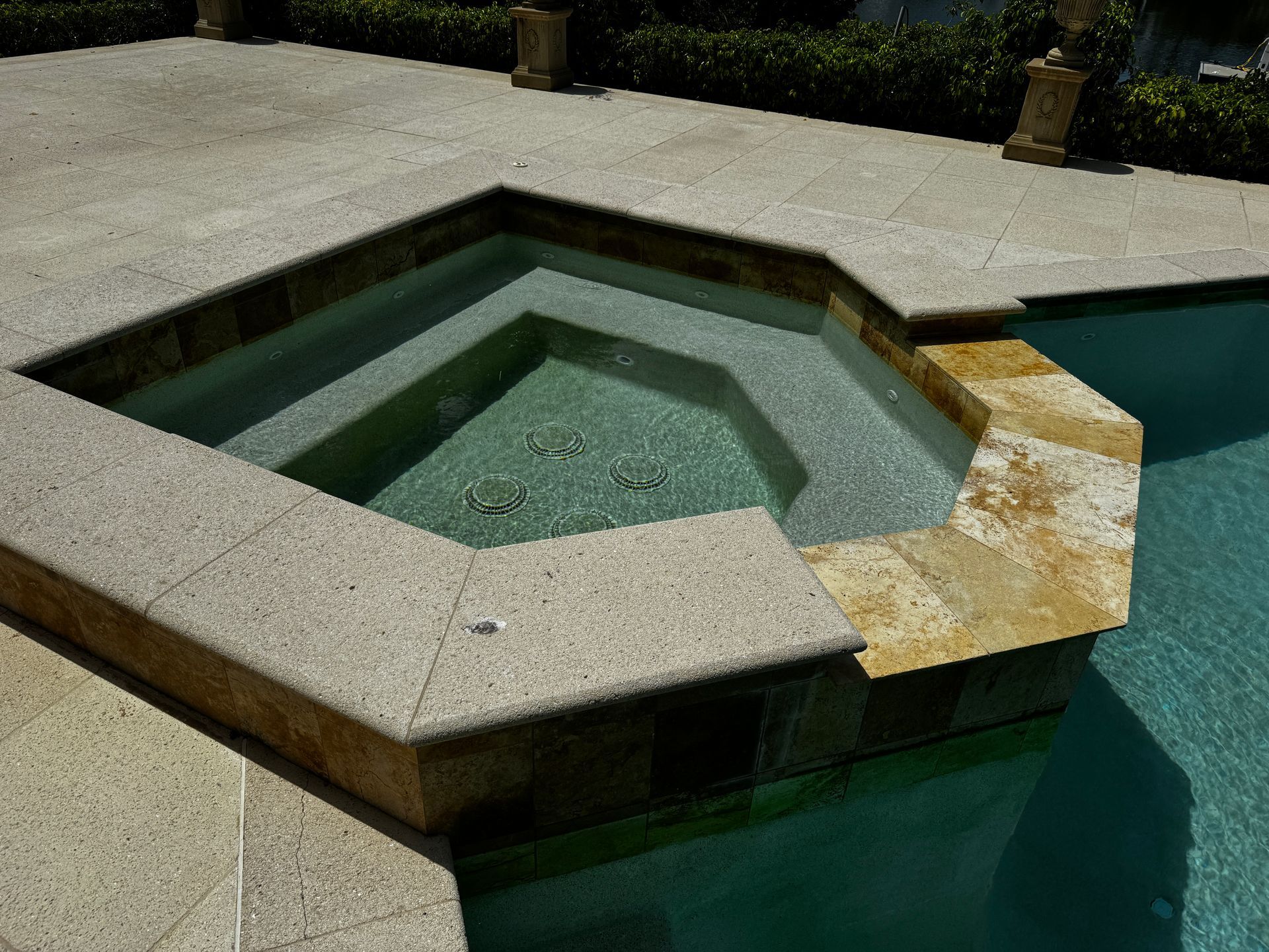 A raised, stone-tiled hot tub sits adjacent to a turquoise swimming pool on a paved patio.