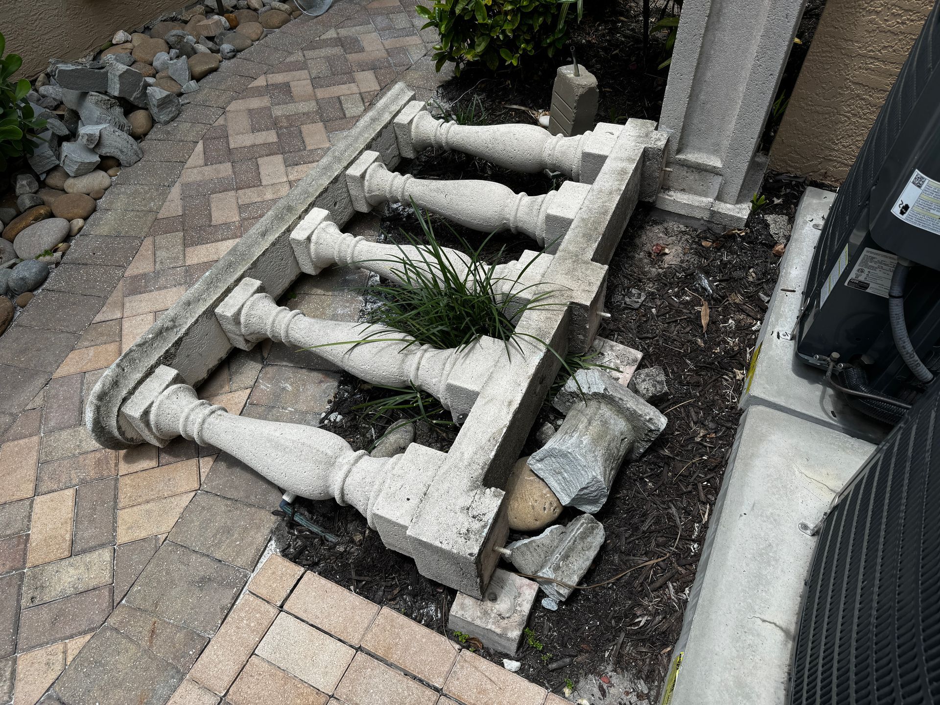 A broken concrete balustrade lying on dark mulch next to a brick walkway and a mechanical unit.