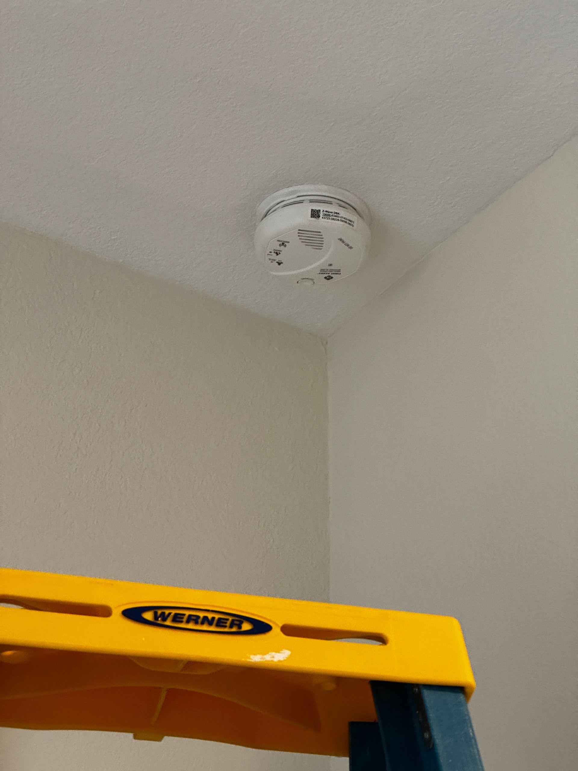 A smoke detector mounted on a white ceiling above a yellow Werner ladder.