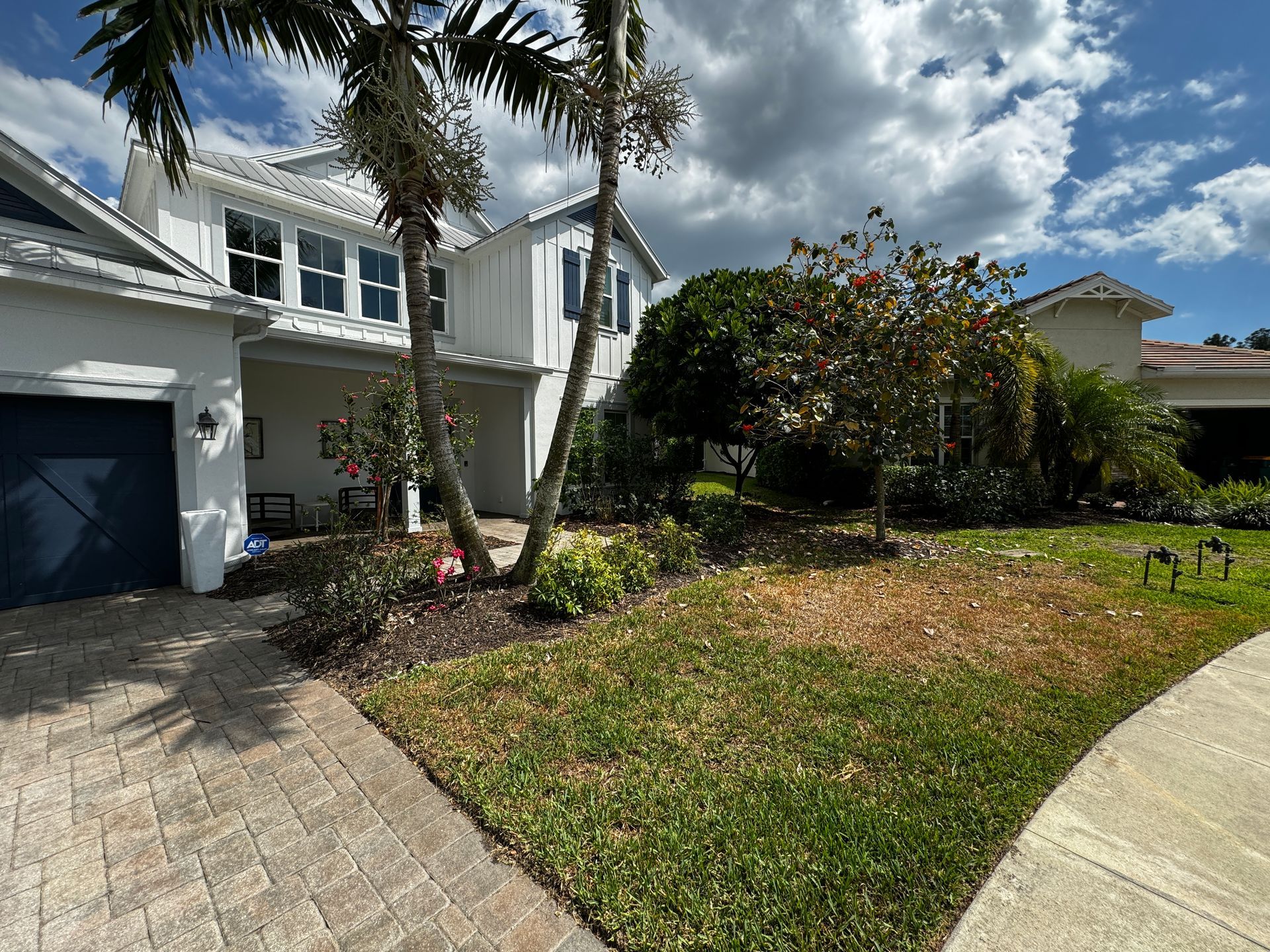 A white two-story house with a blue garage door, palm trees, and a patchy lawn under a partly cloudy sky.