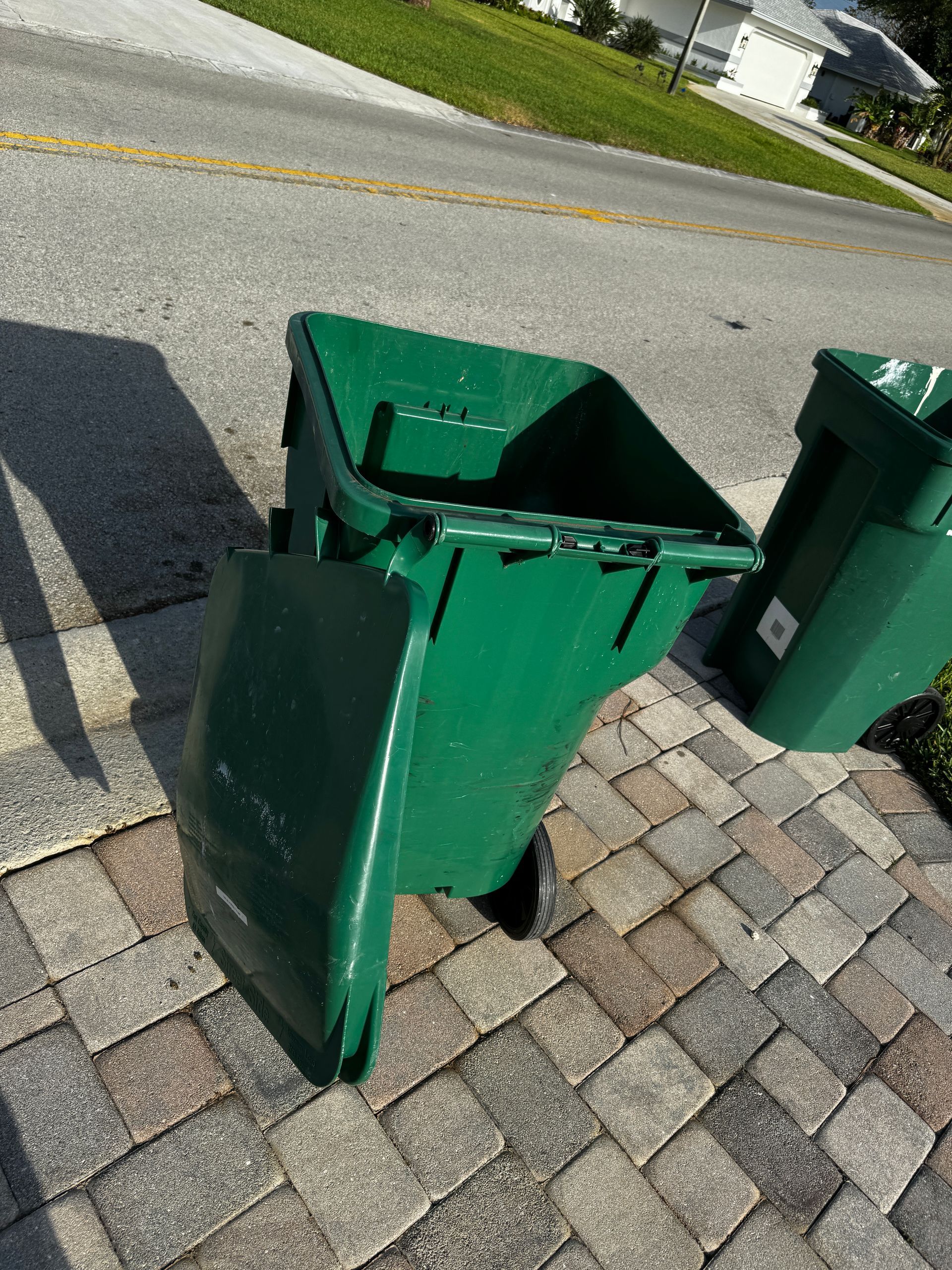 Two green rolling trash bins with lids detached sit on a paved driveway next to a road.
