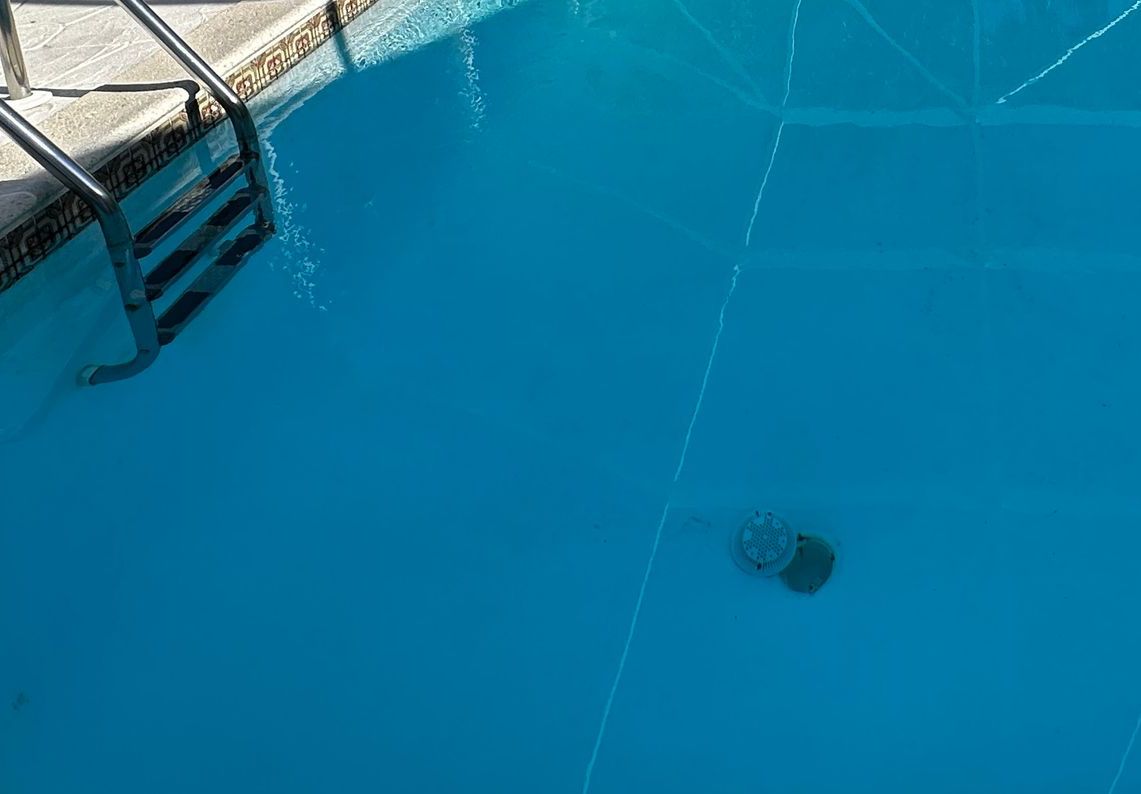 A high-angle view of a bright blue swimming pool with metal ladder steps on the left and a drain cover on the bottom.