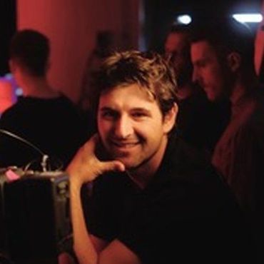 Man smiling, hand near camera. Dim setting with others in the background.