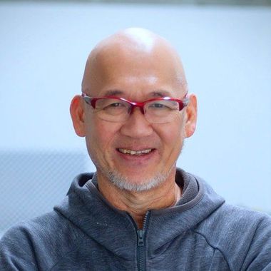 Bald man with glasses wearing a gray hoodie smiles, indoors with a light background.