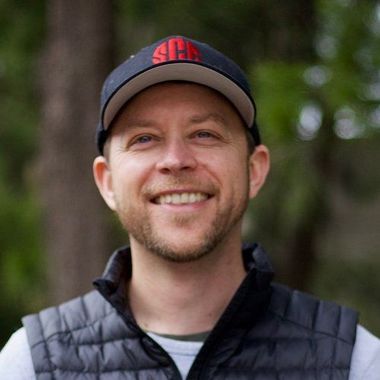 Man wearing a black hat and vest smiles widely, set in front of blurred trees.