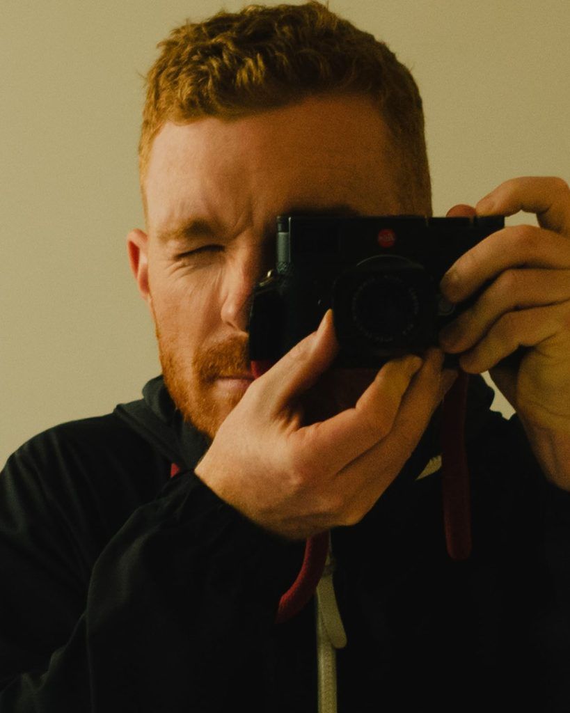 Man with red hair, taking a photo with a black camera. His eye is squinting, indoors.