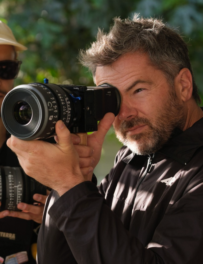 Man looking through a camera lens, outdoors. He wears a dark jacket, has a beard, and focuses intently.